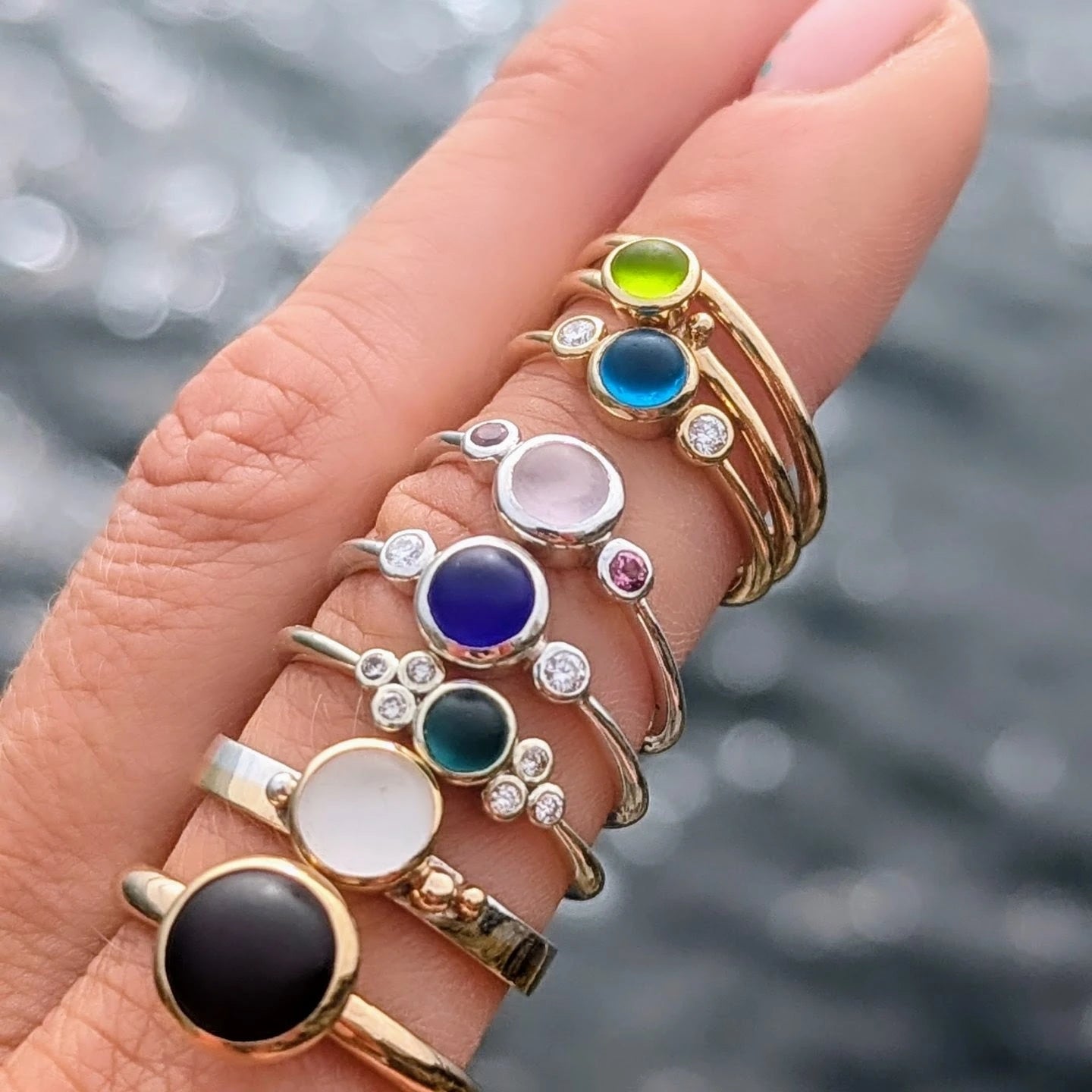 Handmade gold and silver rings with vibrant coloured sea glass, diamonds on a finger above blurred ocean background made by Booblinka Jewellery