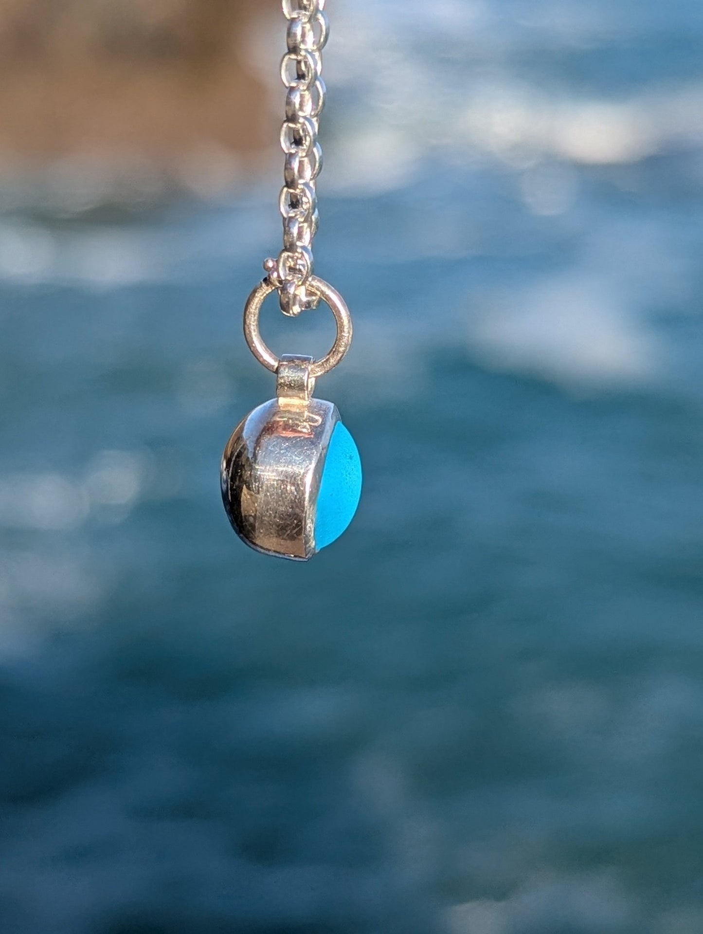 Side detail of round blue sea glass necklace showing rounded back above blurred ocean background