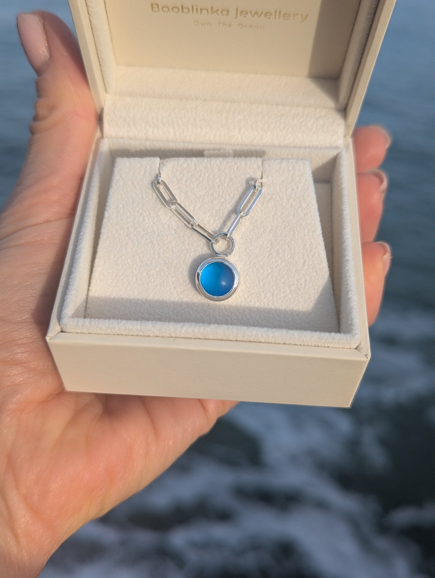 Hand holding an open jewellery box with round blue sea glass silver necklace above the ocean