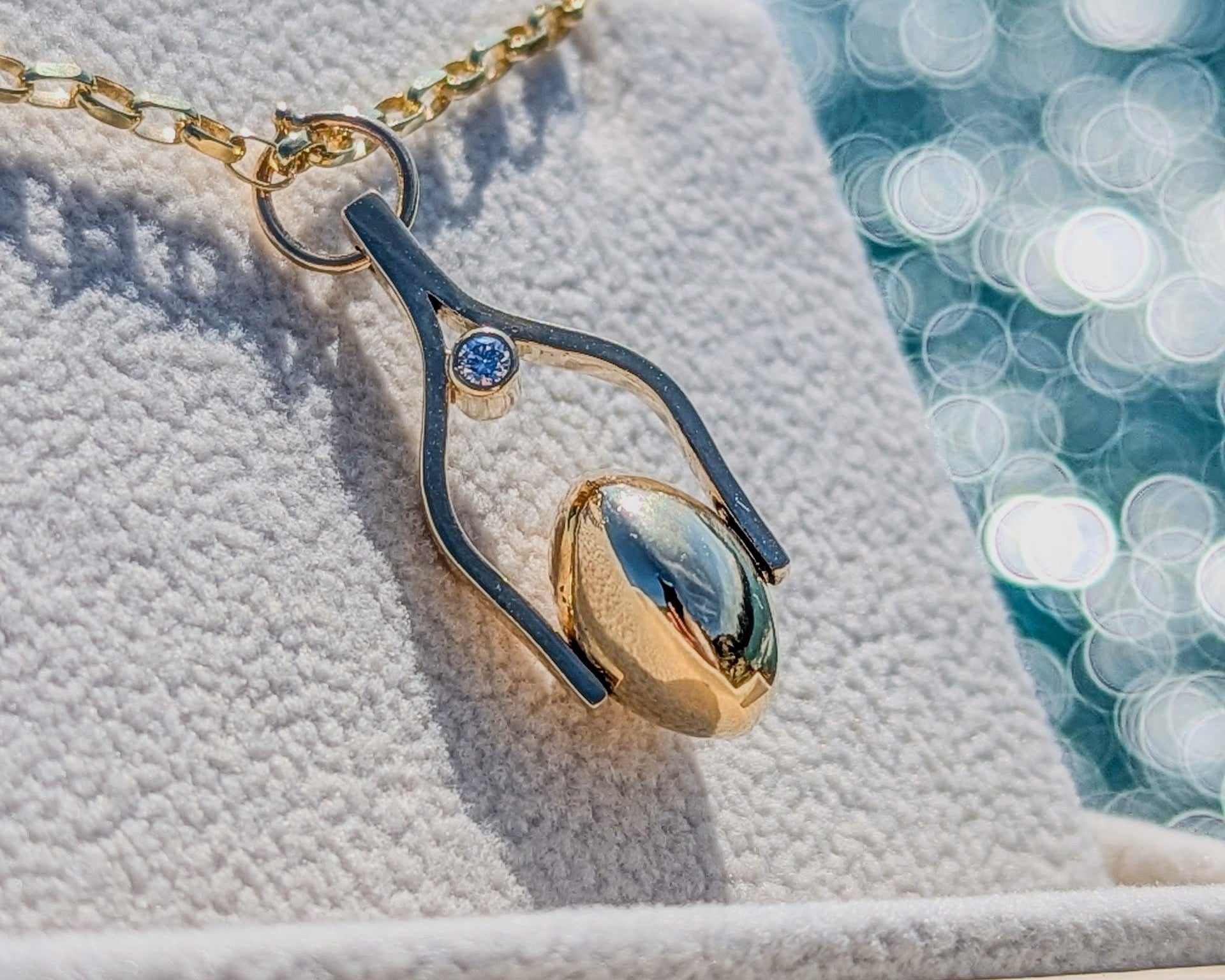 Gold spinning moon pendant with ocean diamonds on a textured surface with a bokah ocean background made by Booblinka Jewellery