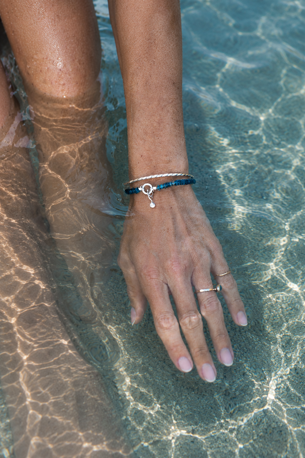 A close-up of a person's hand resting in clear, sunlit pool water, wearing a beaded silver bracelet and a ring.