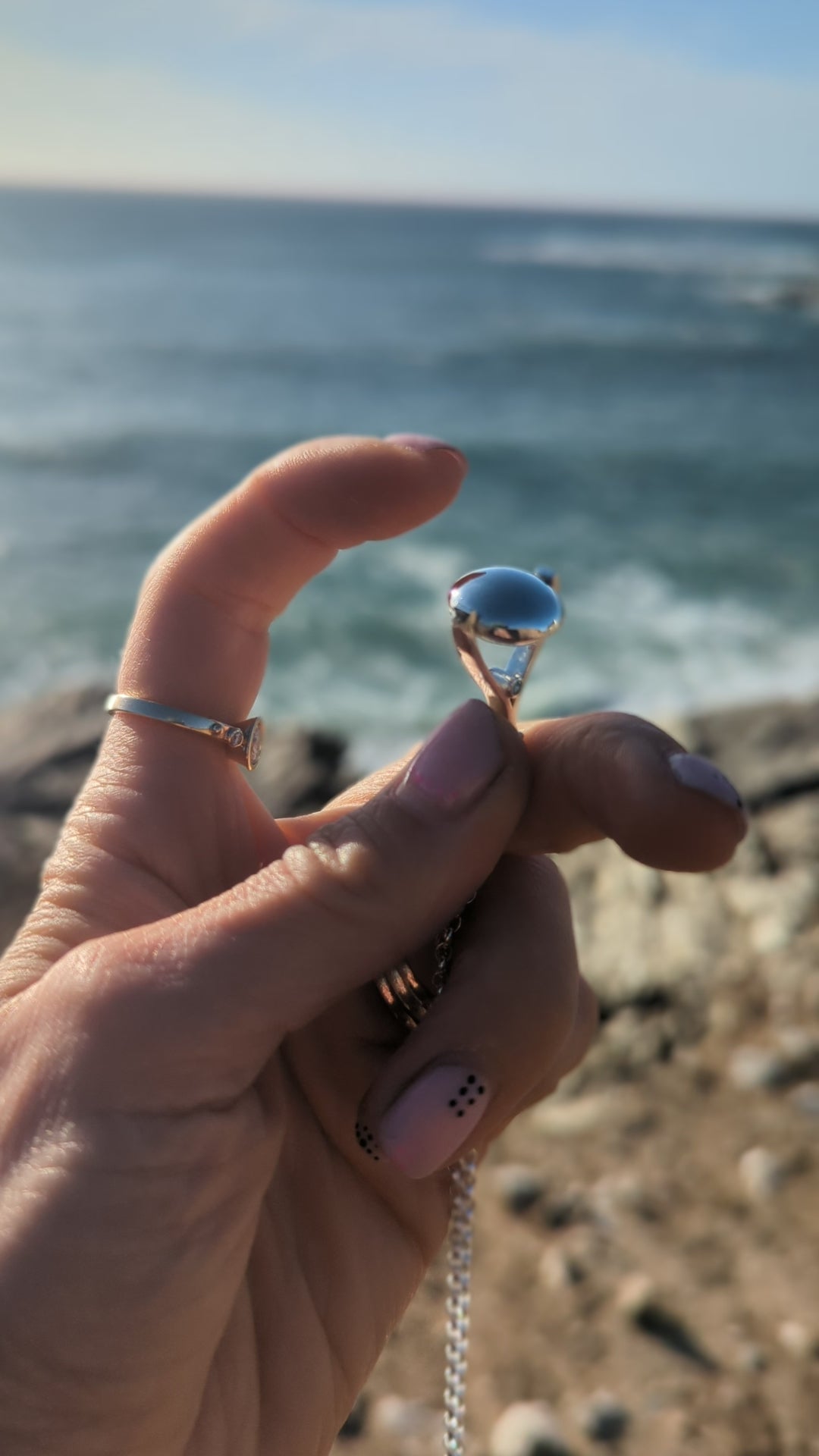 Hand holding silver spinning necklace with ocean background, showing the spinning movement. By Booblinka Jewellery.