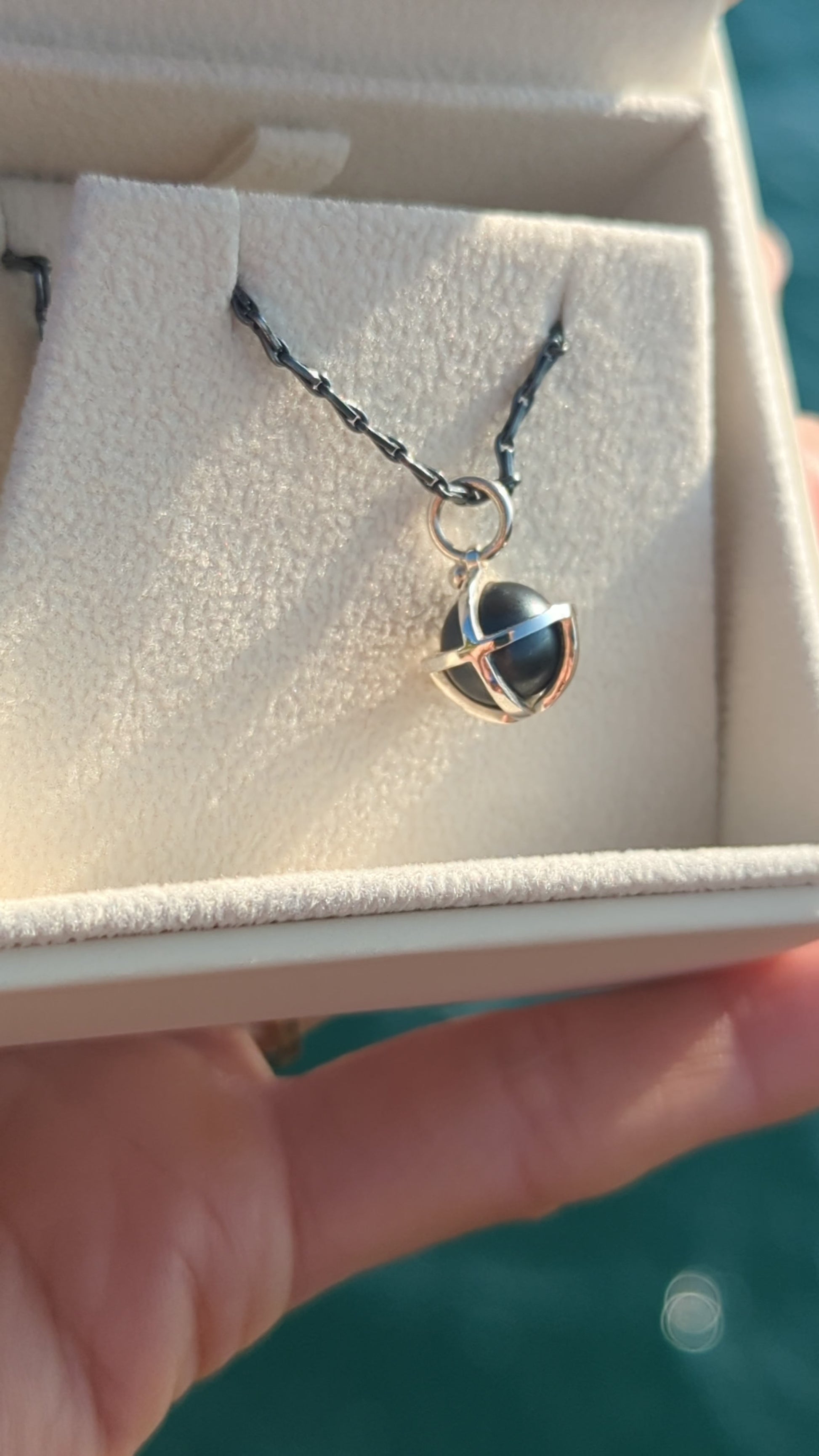 Video of black hematite sphere in silver cage necklace in plush ivory jewellery box hold by hand above turquoise ocean. Made by Booblinka Jewellery