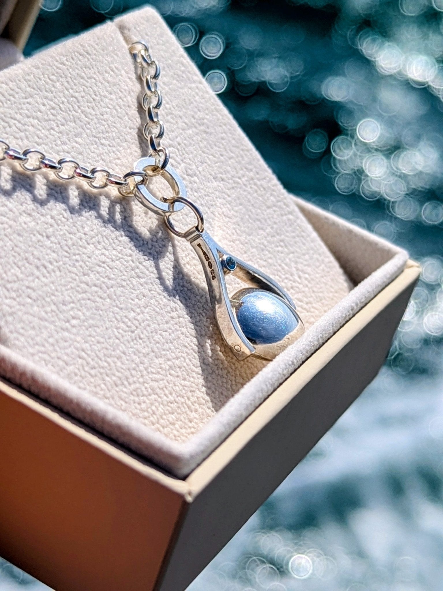 A handcrafted silver spinning necklace featuring a London blue topaz gemstone, resting in a jewellery box with the sea sparkling in the background made by Booblinka Jewellery.