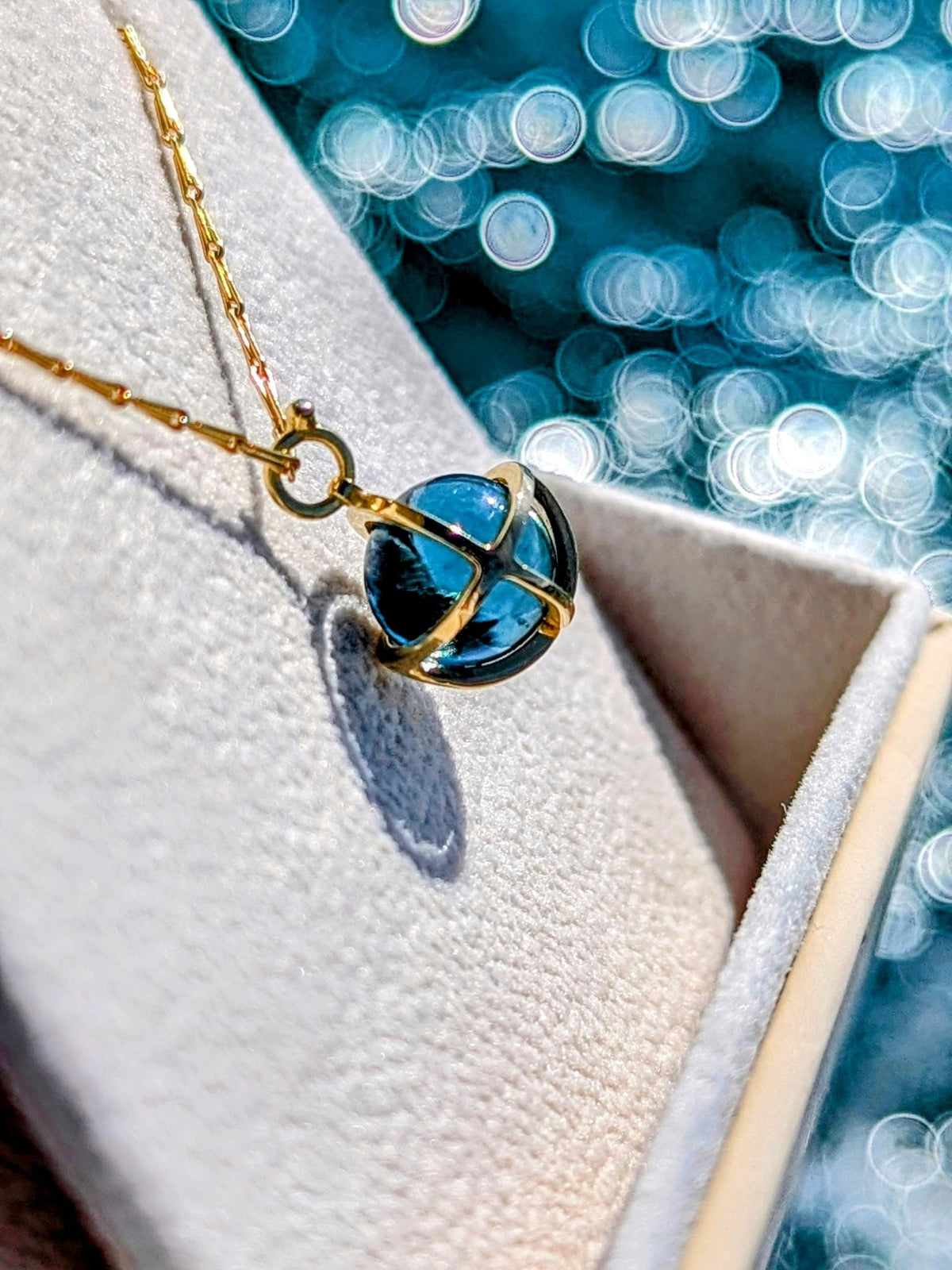 18 Carat Gold Necklace with London Blue Topaz Sphere in gold cage in a jewellery box above shimmering ocean background made by Booblinka Jewellery