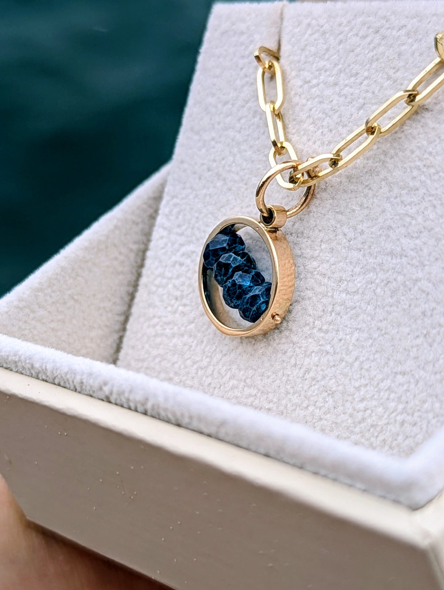 Top view of solid gold blue topaz necklace with paperclip chain in an open ivory jewellery box against deep blue sea.