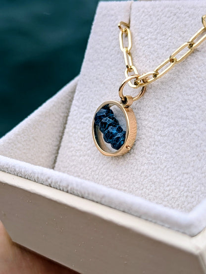 Top view of solid gold blue topaz necklace with paperclip chain in an open ivory jewellery box against deep blue sea.