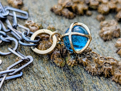 Swiss Blue Topaz sphere pendant with a solid 9ct gold hinged charm holder resting on coastal rocks beside a black rhodium sterling silver chain.
