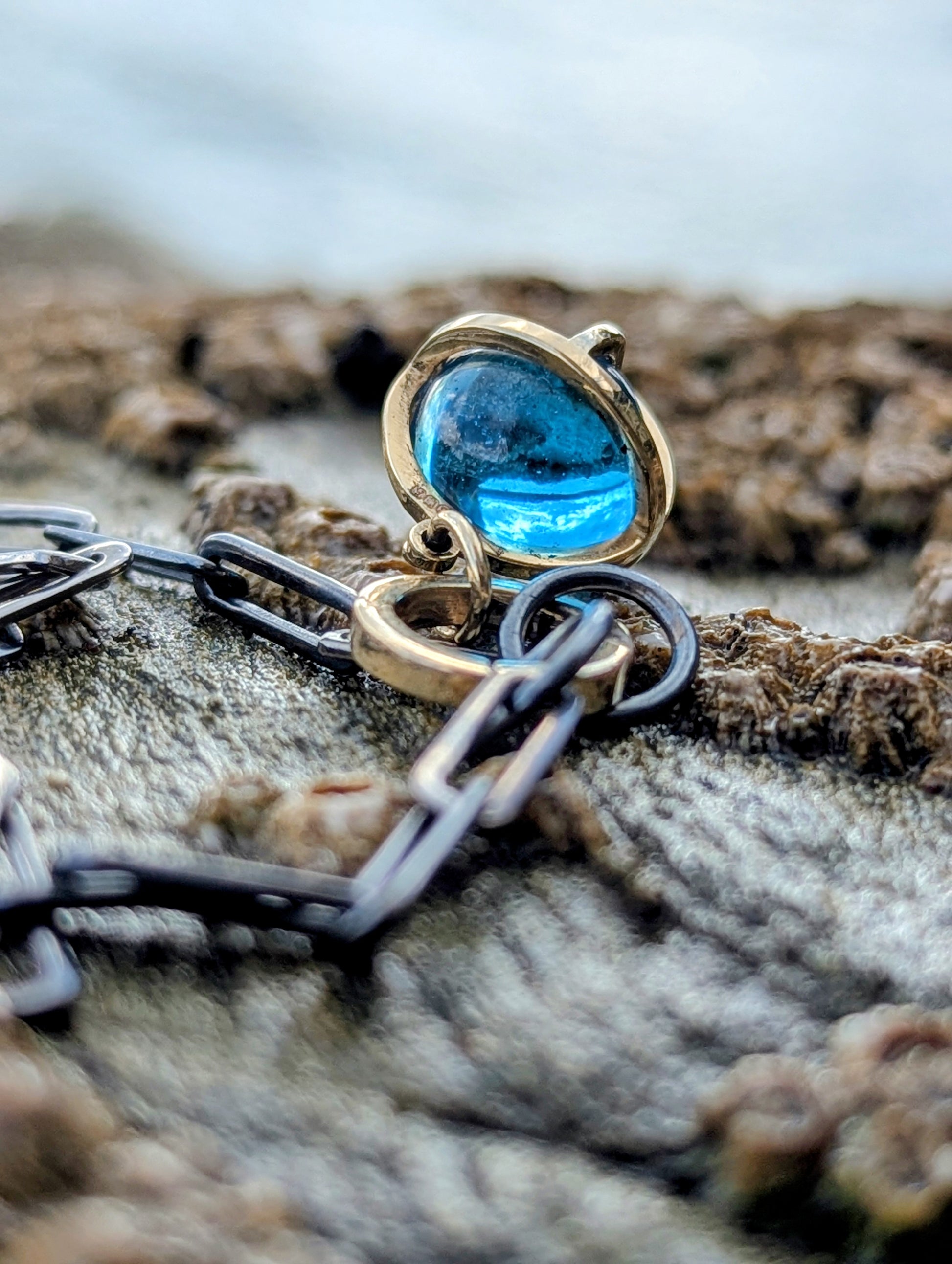 Close-up of a handmade 9ct gold Swiss Blue Topaz sphere pendant resting on coastal rocks, attached to a black rhodium sterling silver chain.