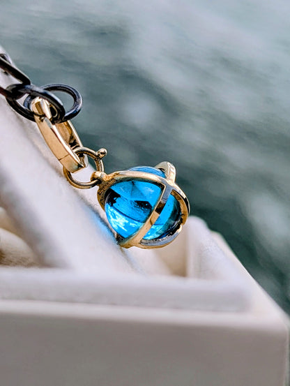 Close-up of a Swiss Blue Topaz sphere necklace in a solid 9ct gold cage hanging from a black rhodium sterling silver chain.