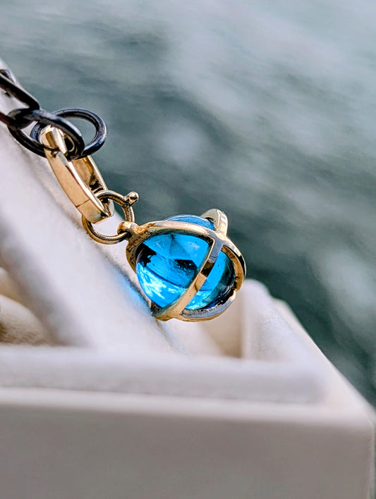 Close-up of a Swiss Blue Topaz sphere necklace in a solid 9ct gold cage hanging from a black rhodium sterling silver chain.