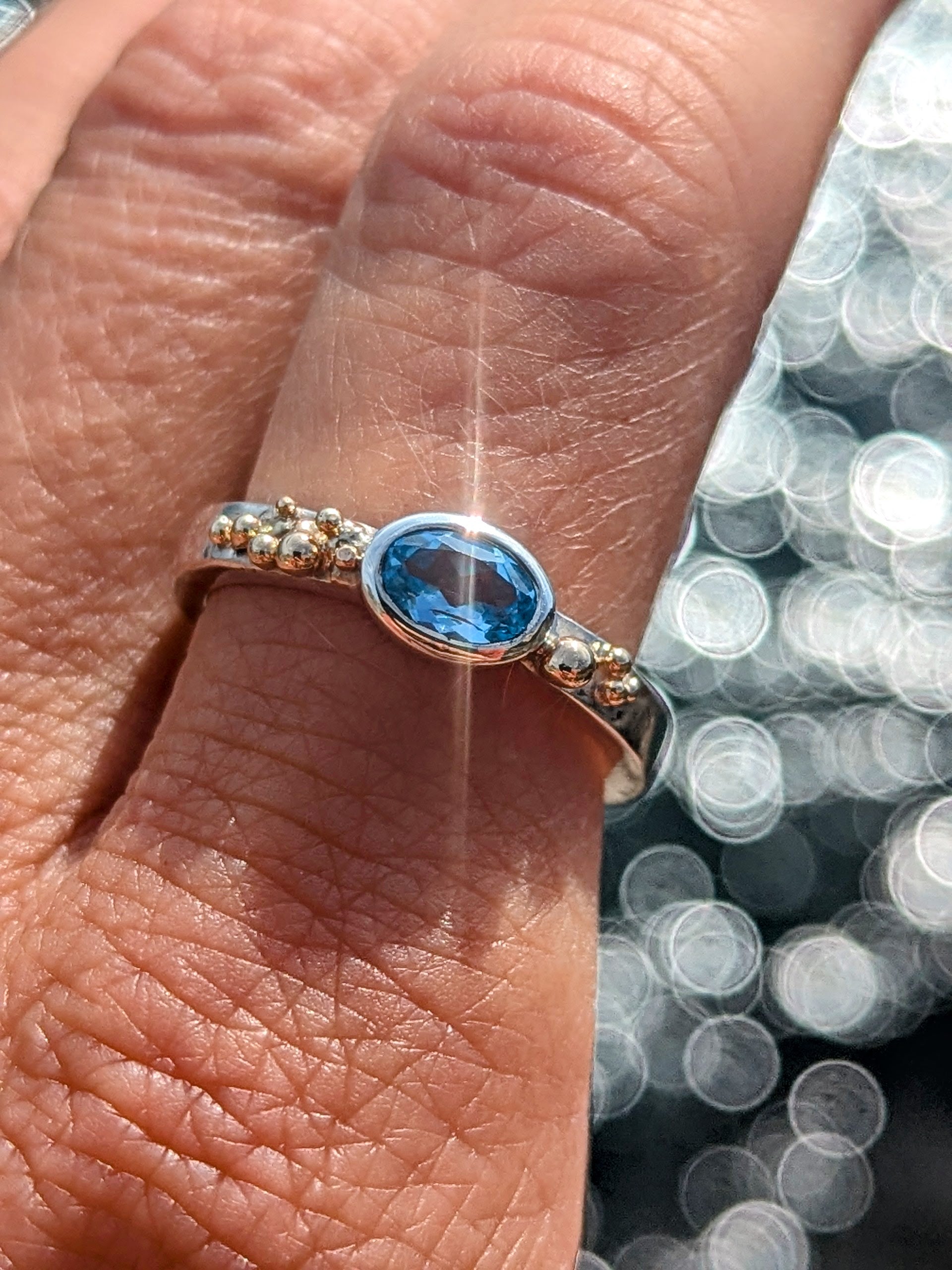 Silver ring with a blue topaz and gold granules on a finger against a blurred ocean background made by Booblinka Jewellery