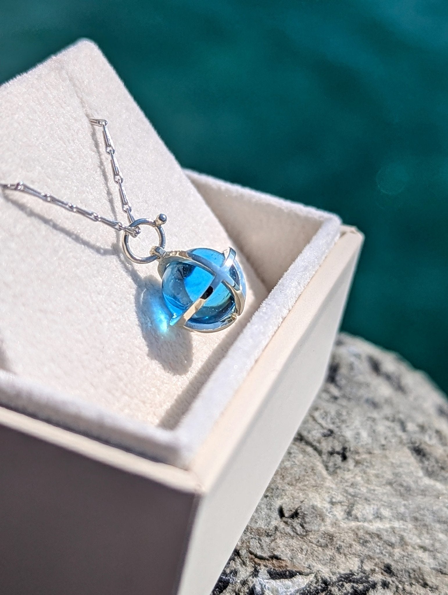 Blue topaz sphere in white gold cage necklace in a jewellery box with ocean background made by Booblinka Jewellery