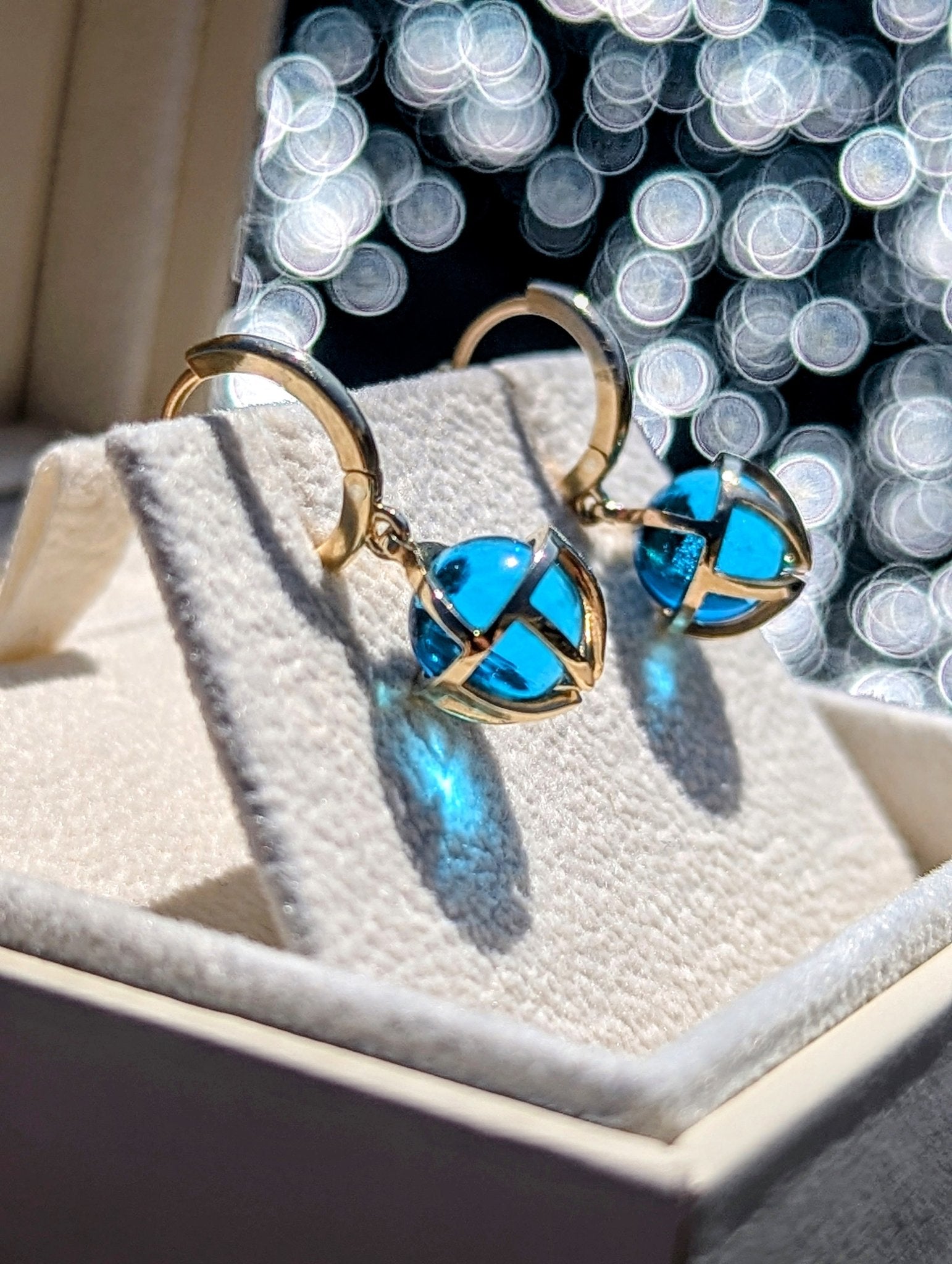 Gold hoop earrings with blue topaz spheres in gold cages in a jewellery box with a shimmering ocean in a background made by Booblinka Jewellery