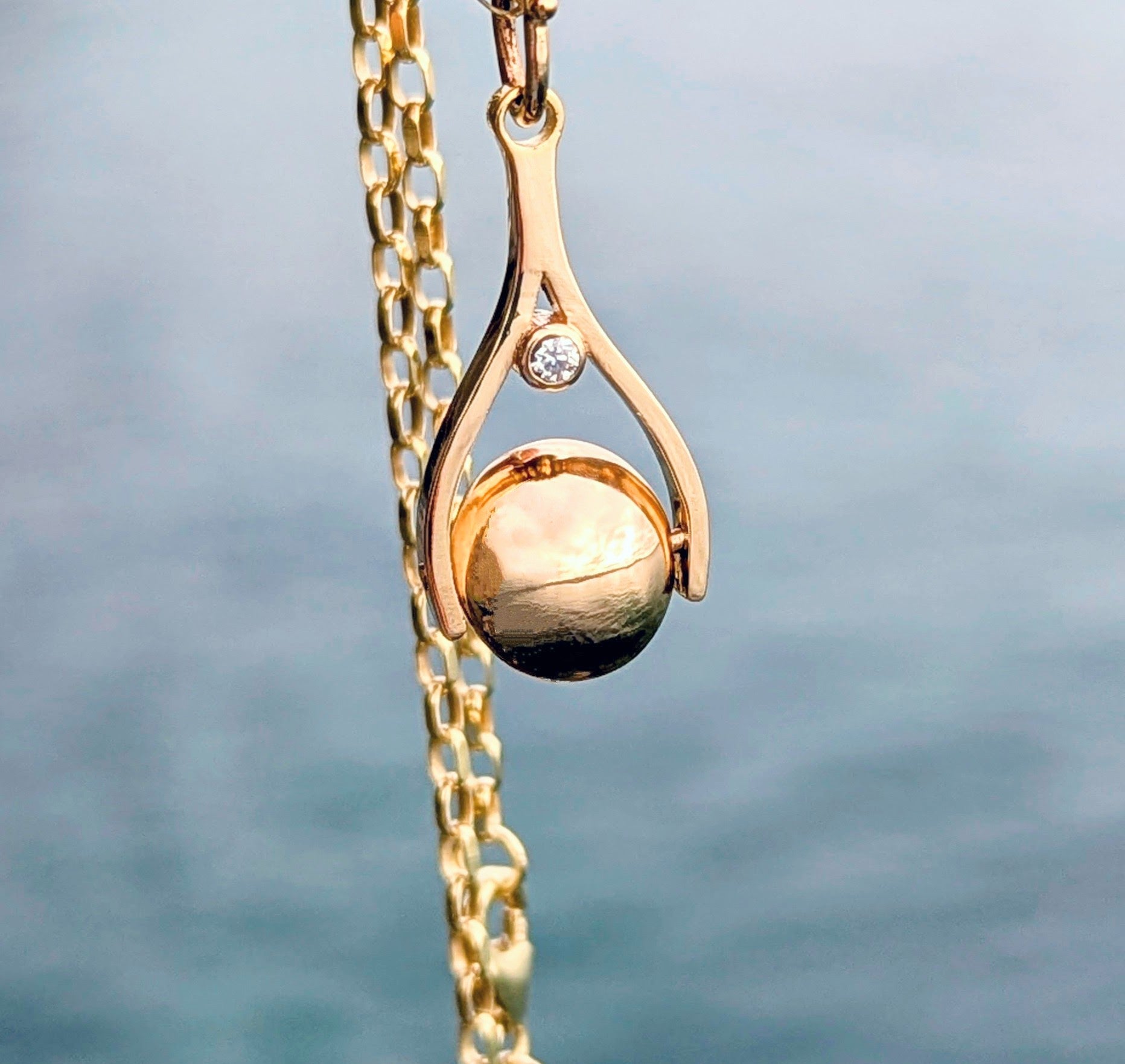 Gold necklace with a spinning pendant and diamond against a blurred ocean background made by Booblinka Jewellery