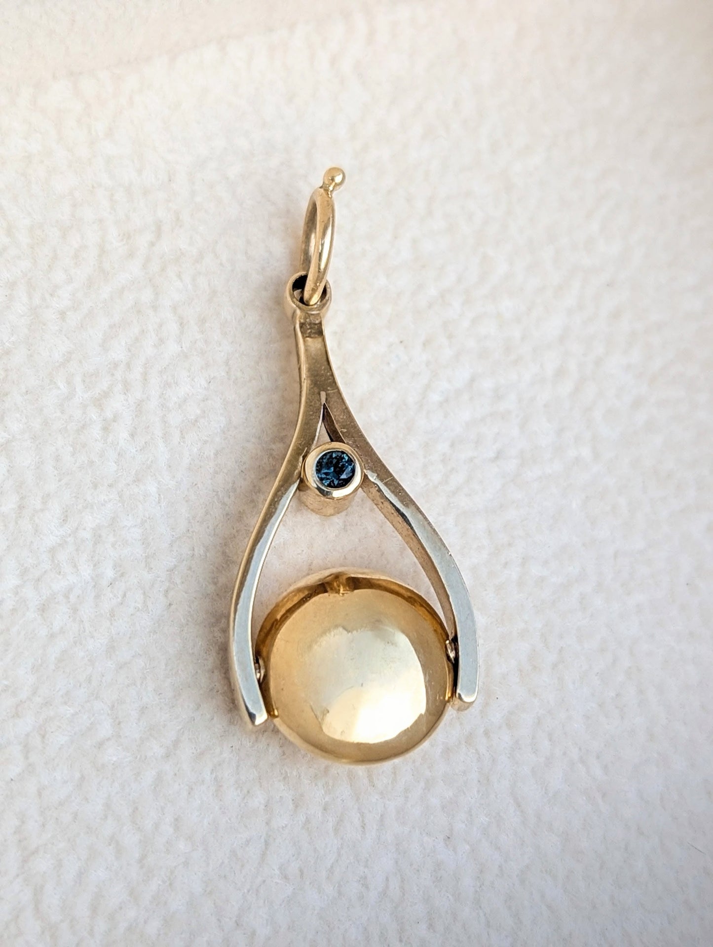 Gold pendant with a teal sapphire and spinning bead on a textured white background made by Booblinka Jewellery