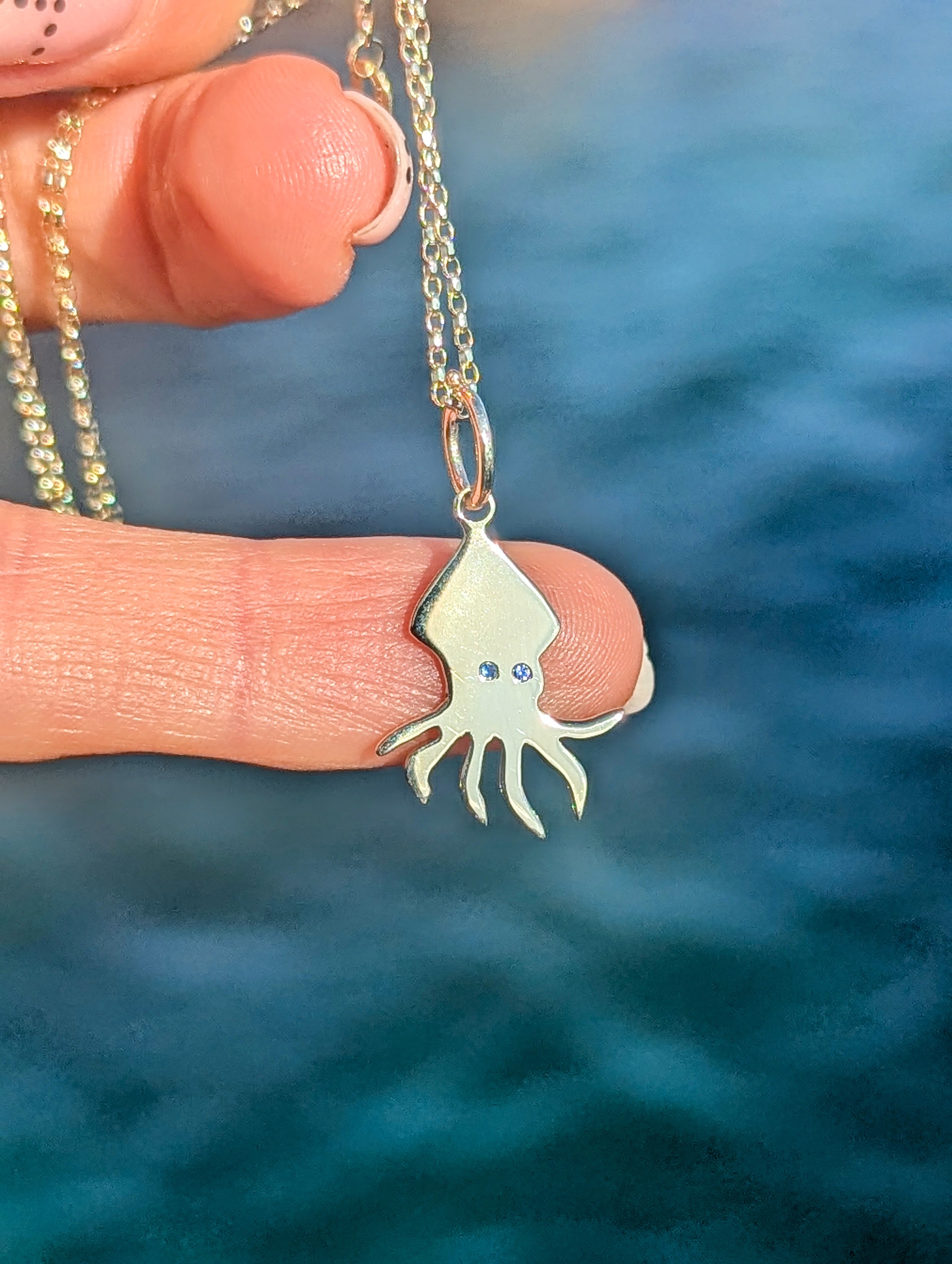 Gold squid pendant with blue sapphire eyes on a chain held between fingers against an ocean background made by Booblinka Jewellery.