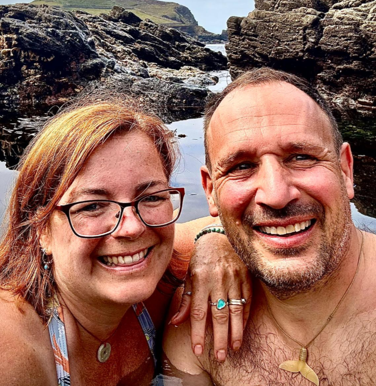 Two people smiling at the camera with a rocky coastal background wearing handmade gold sea glass anniversary ring by Booblinka Jewellery