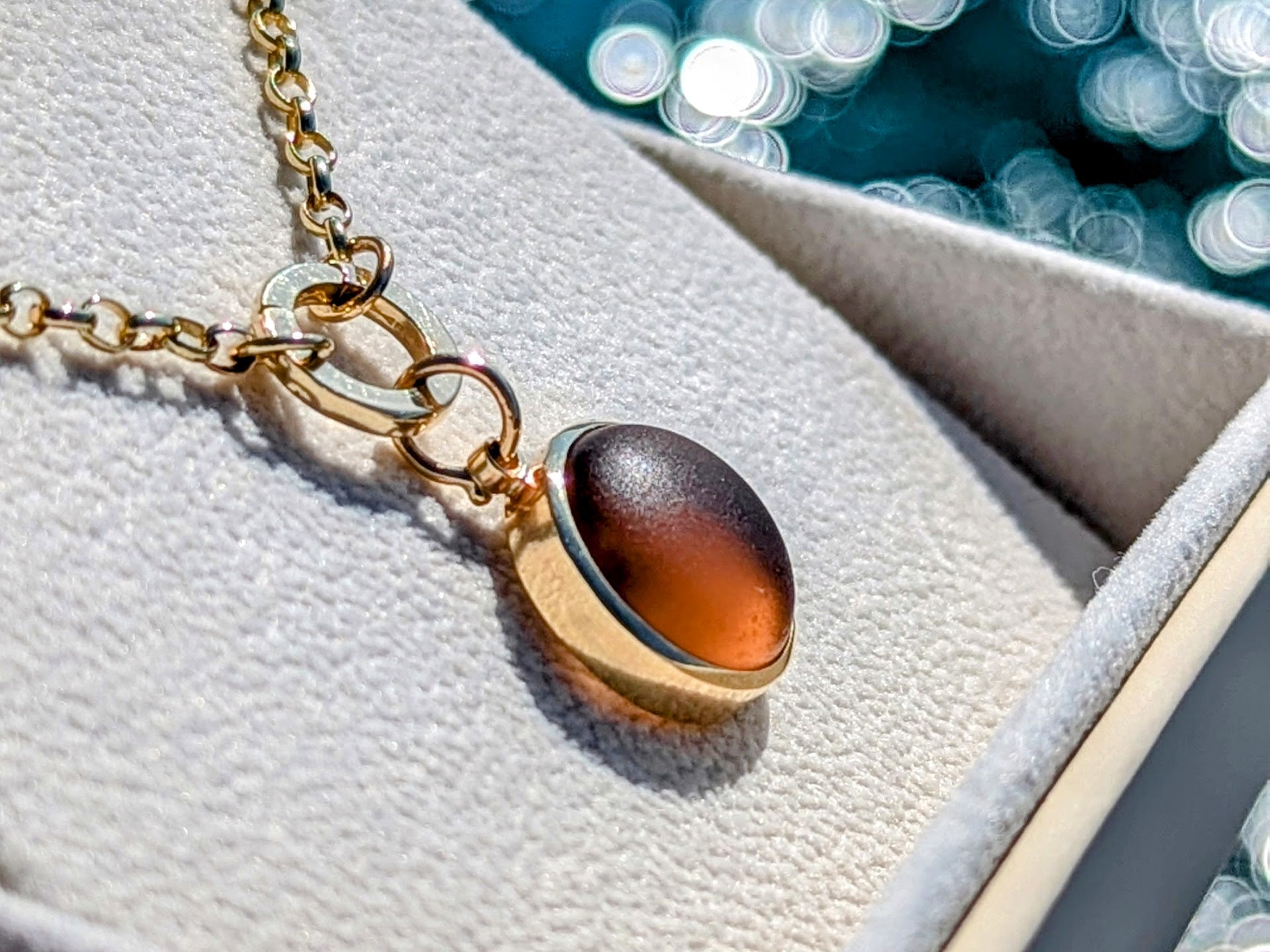 Brown sea glass gold necklace in an ivory plush jewellery box above shimmering ocean handmade by Booblinka Jewellery