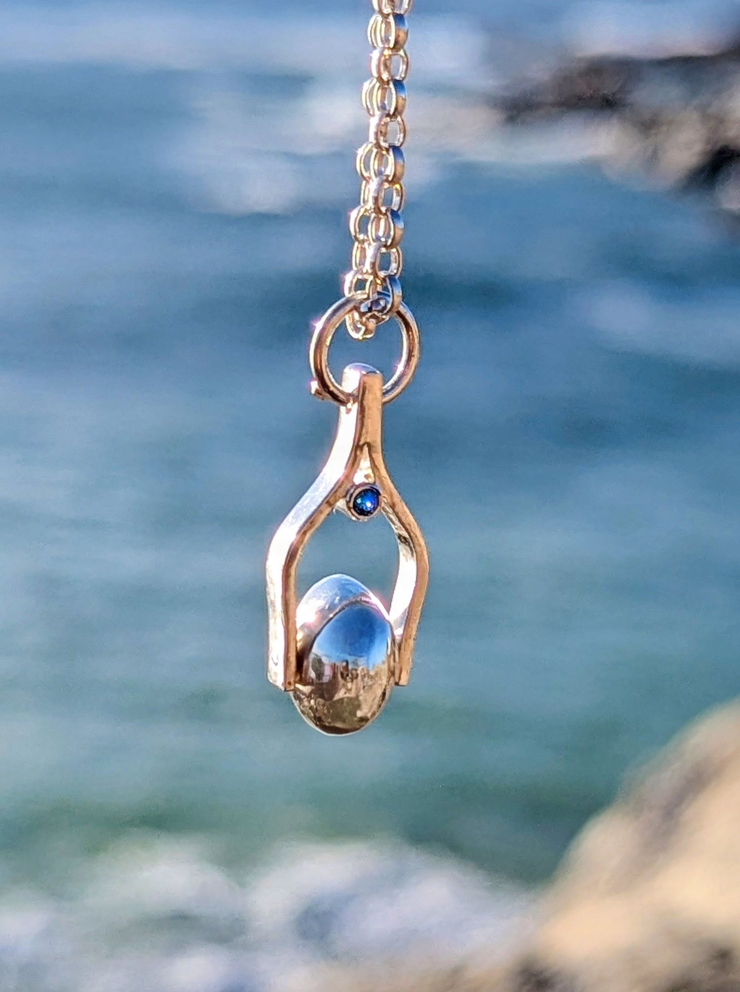 Silver necklace with a spinning pendant with a blue topaz against a blurred ocean background