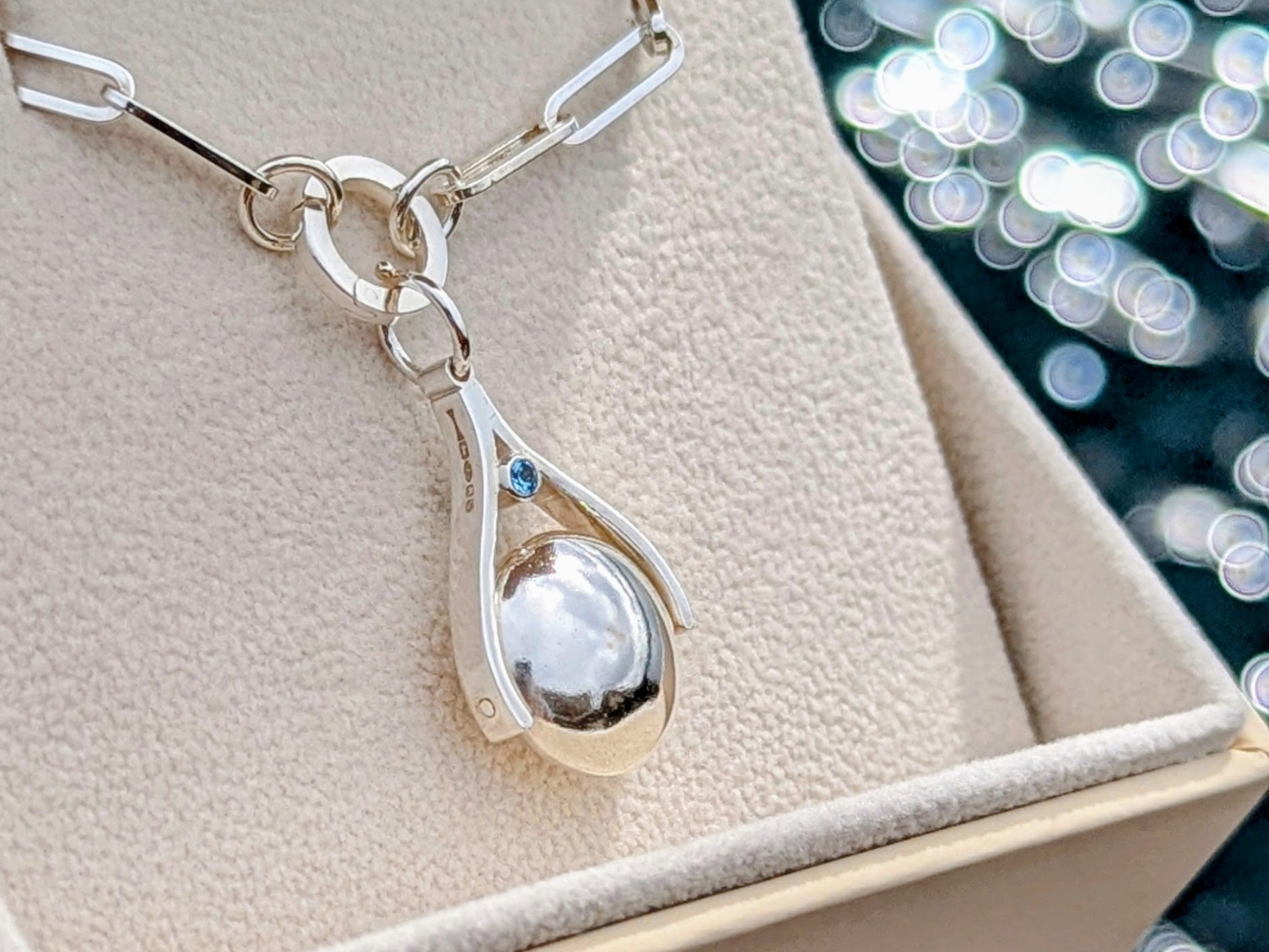 Silver spinning pendant with blue topaz on a paperclip chain in a jewellery box against blurred ocean background made by Booblinka Jewellery.