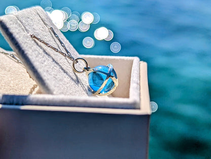 Jewellery box with a blue topaz sphere pendant necklace made in white gold against a blurred water background by Booblinka Jewellery