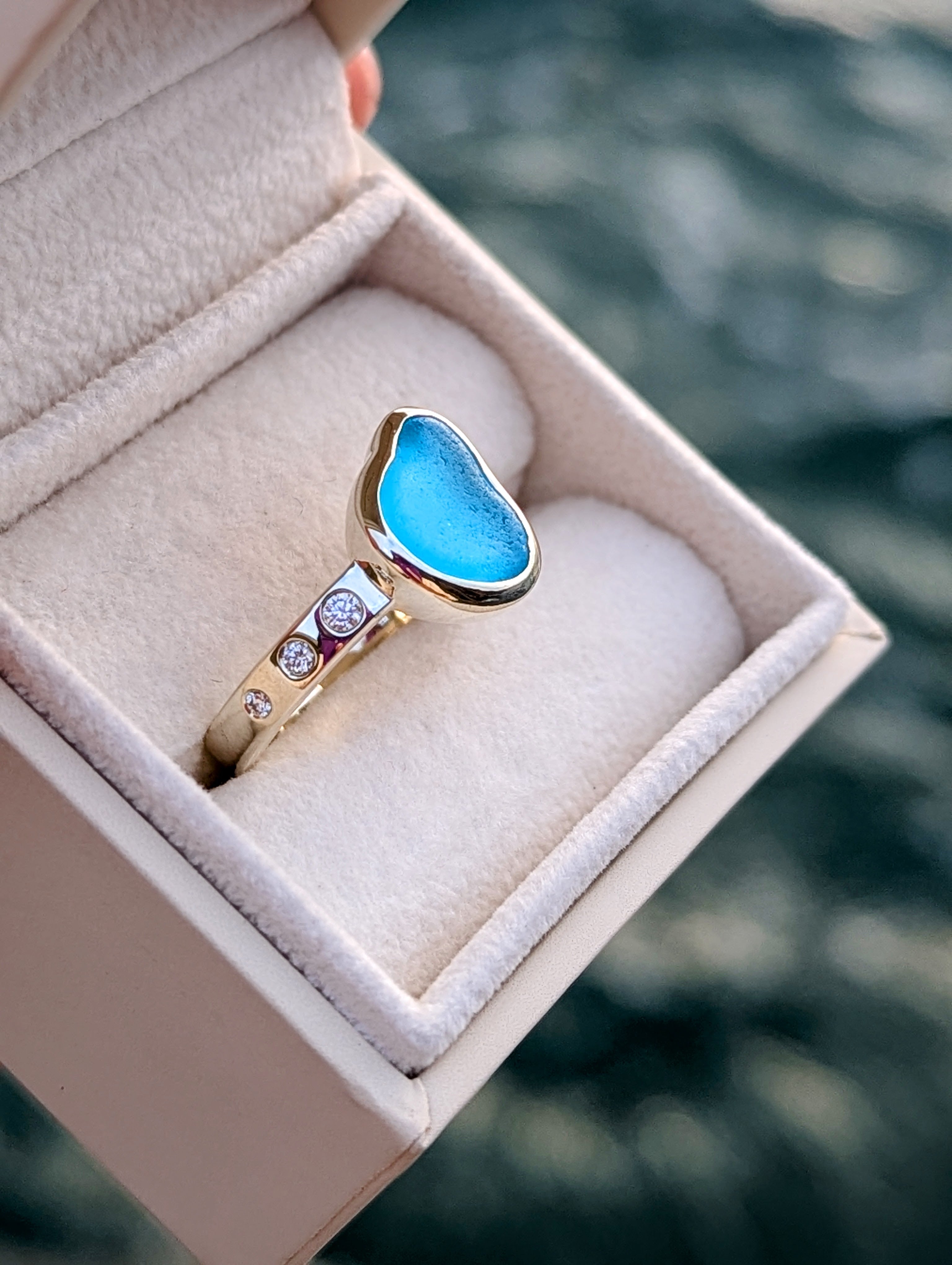 White gold Ring with blue sea glass, diamonds in a jewellery box against a blurred ocean background made by Booblinka Jewellery