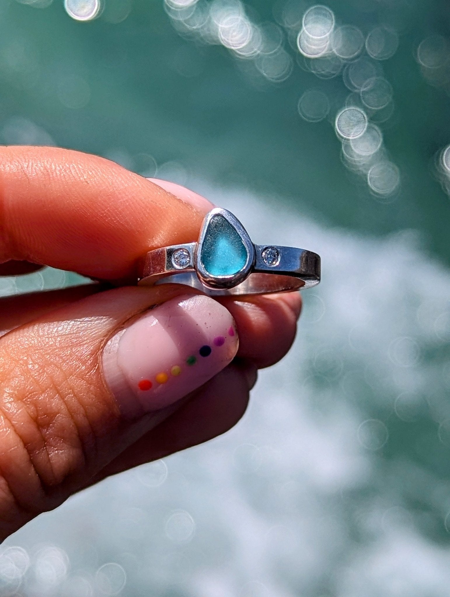 Hand holding Aqua Blue Sea Glass and Moissanite Silver Ring  from Allure Collection above shimmering ocean by Booblinka Jewellery