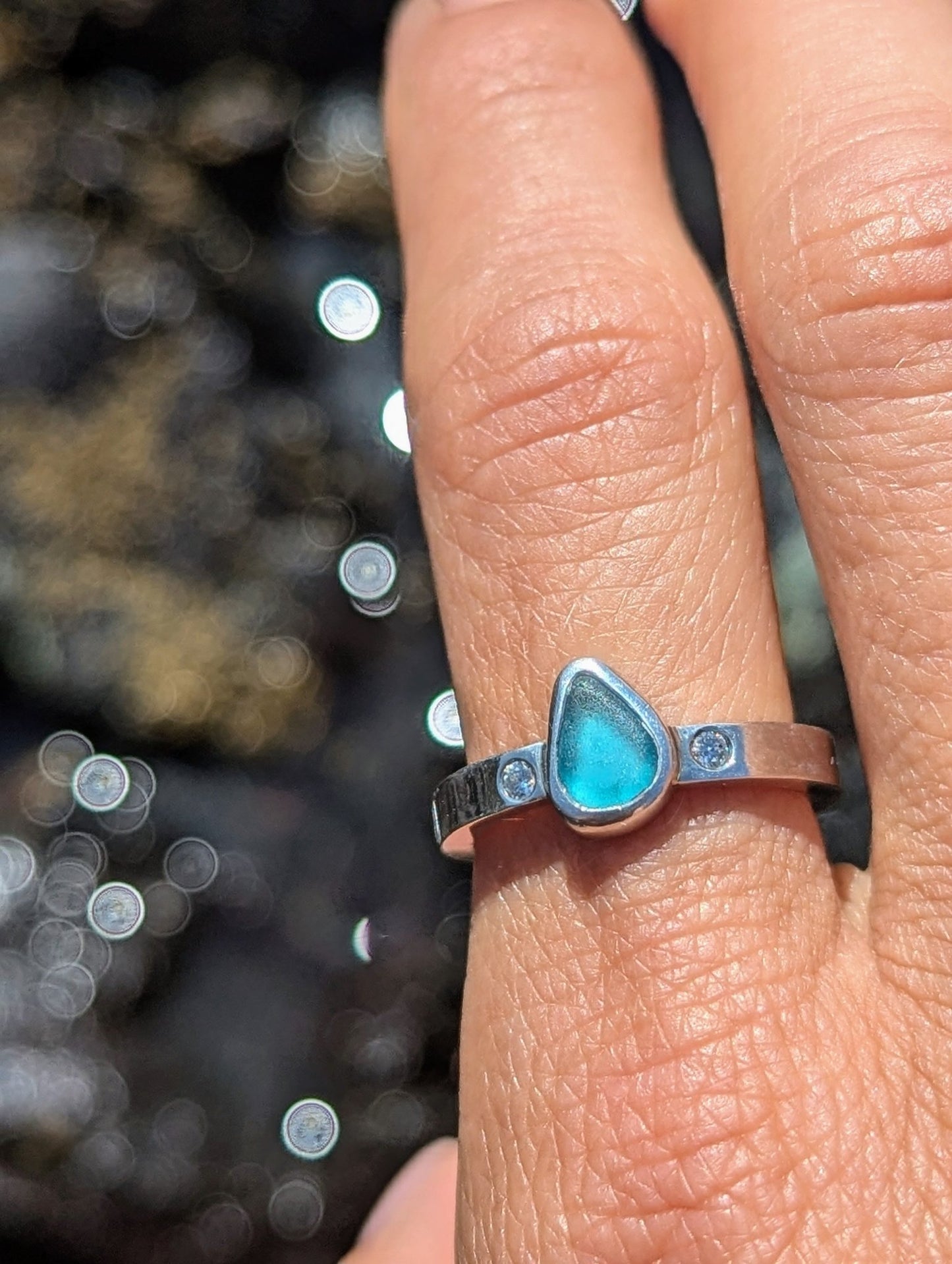 Aqua Blue Sea Glass and Moissanite Silver Ring  from Allure Collection on  a finger above sparkling water by Booblinka Jewellery