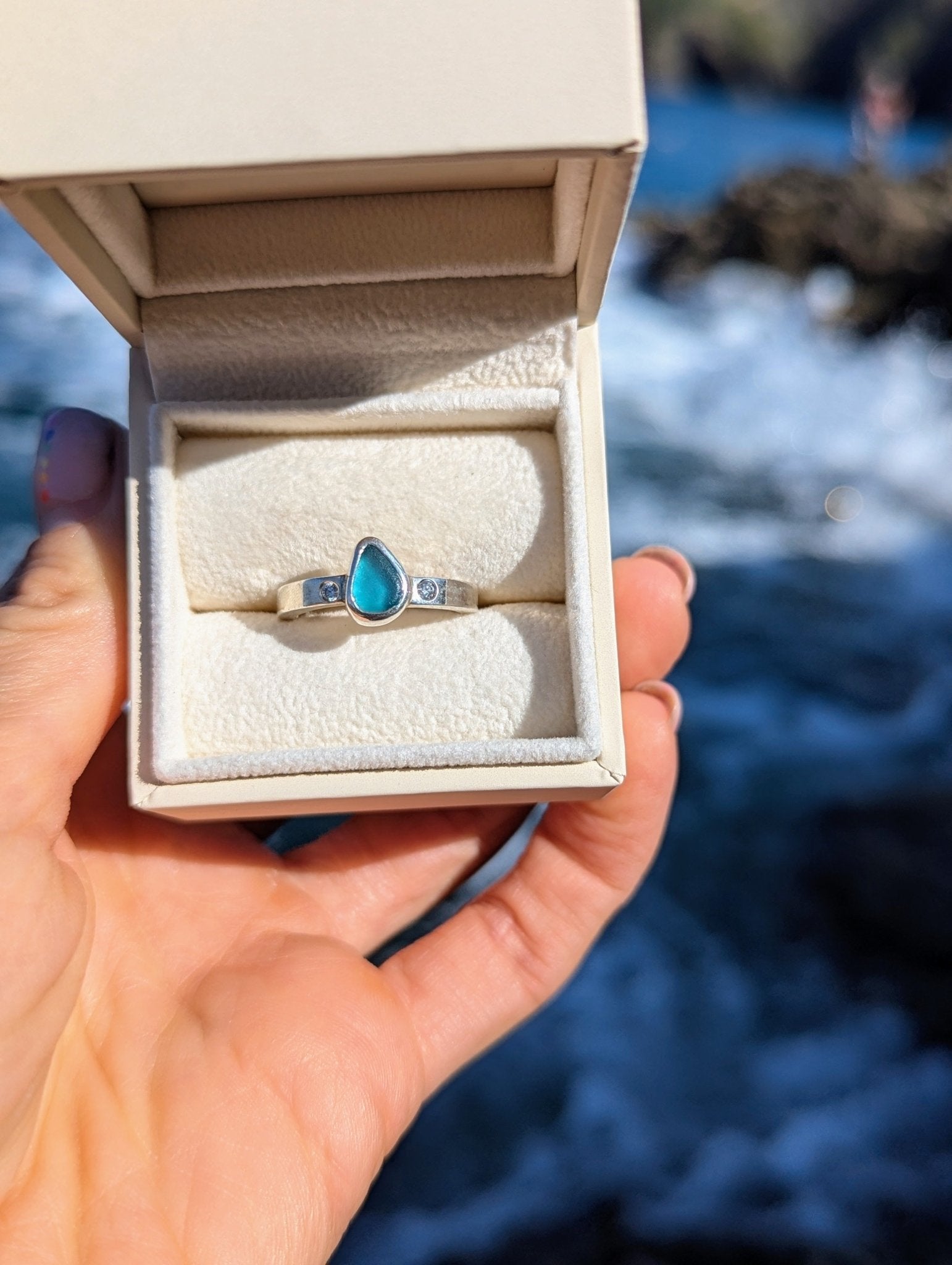 Aqua Blue Sea Glass and Moissanite Silver Ring  from Allure Collection in an ivory plush jewellery box held above the ocean by Booblinka Jewellery 