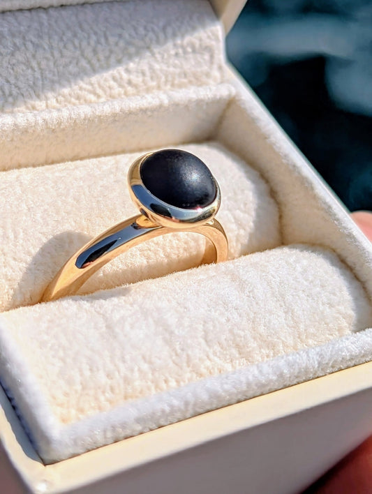 Rare opaque black sea glass ring handmade in solid 9ct yellow gold, displayed in a jewellery box against ocean background