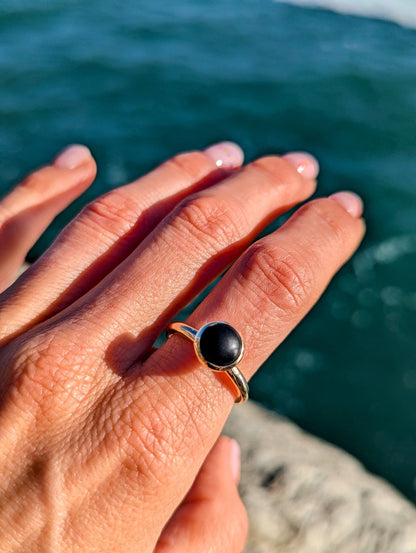 Rare opaque black sea glass and solid gold ring held in the hand with the ocean in the background