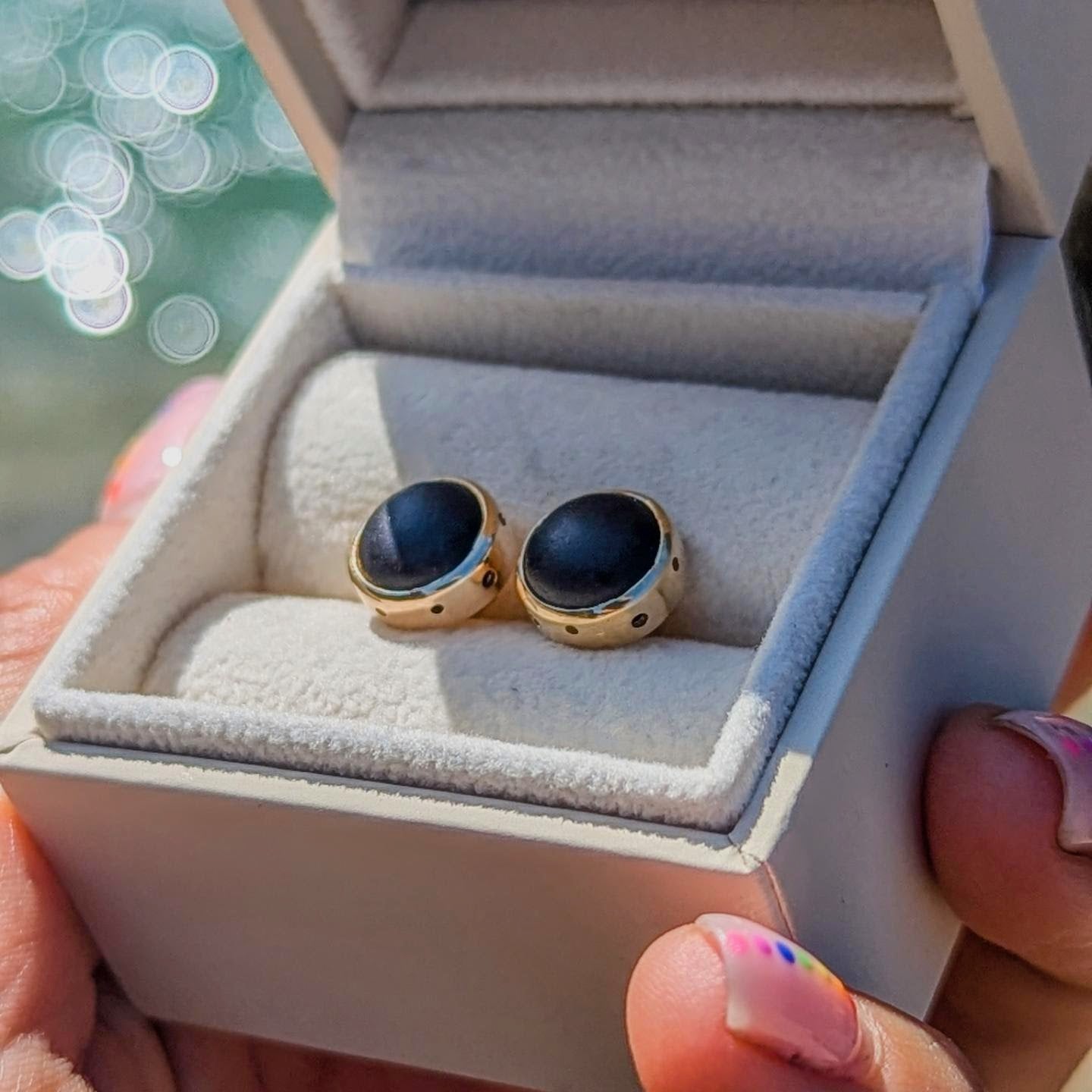 Pair of rare opaque black sea glass stud earrings handmade in solid 9ct yellow gold, displayed in a jewellery box beside the sea.