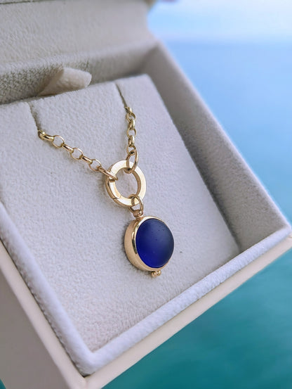 Round blue sea glass pendant set in solid 9ct gold hanging from a solid gold chain with a circular hinged charm holder, displayed in a cream jewellery box.