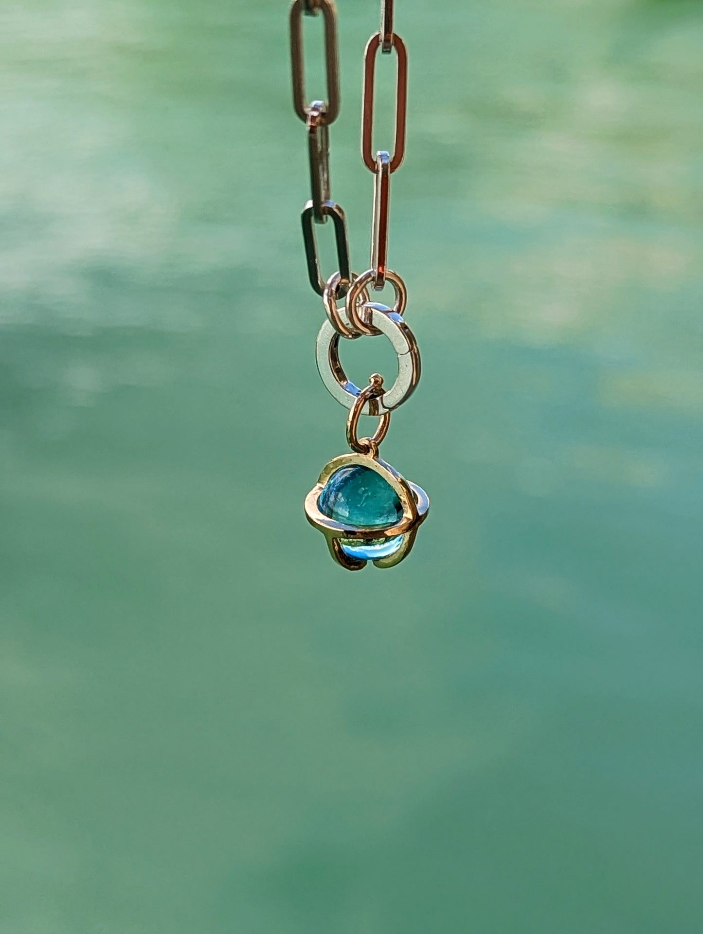 Blue topaz charm hanging from a sterling silver paperclip bracelet with a handmade 9ct gold cage against an ocean background.