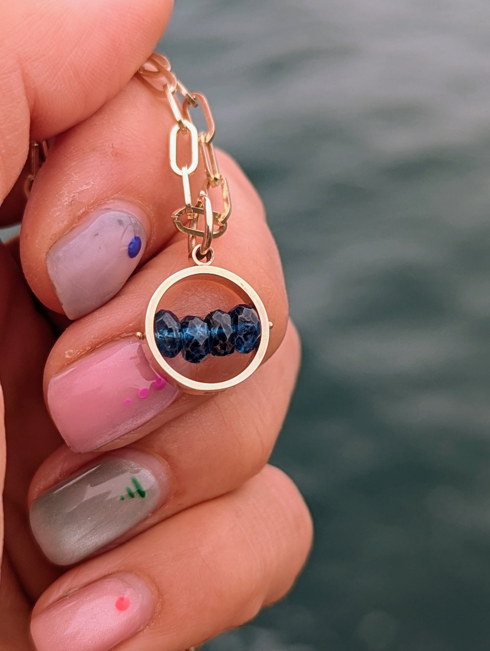 Small circular gold HALO pendant held between fingers, showing four faceted blue topaz gemstones that can be turned by hand.