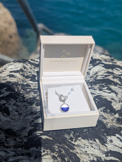 Handmade blue and white sea glass necklace displayed in a jewellery box with a sterling silver paperclip chain on a rocky surface with water in the background