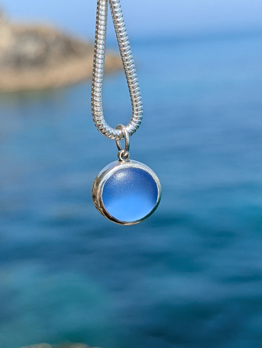 Close-up of a one-of-a-kind blue sea glass necklace handmade in sterling silver with a blurred ocean background