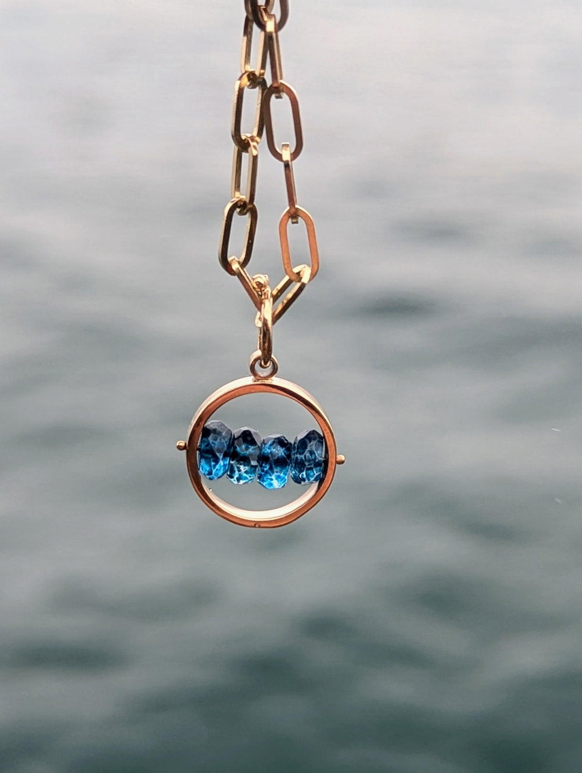 Solid 9ct gold HALO necklace with four faceted blue topaz gemstones across the centre, hanging against the sea.