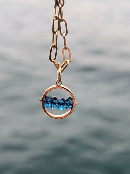 Solid 9ct gold HALO necklace with four faceted blue topaz gemstones across the centre, hanging against the sea.