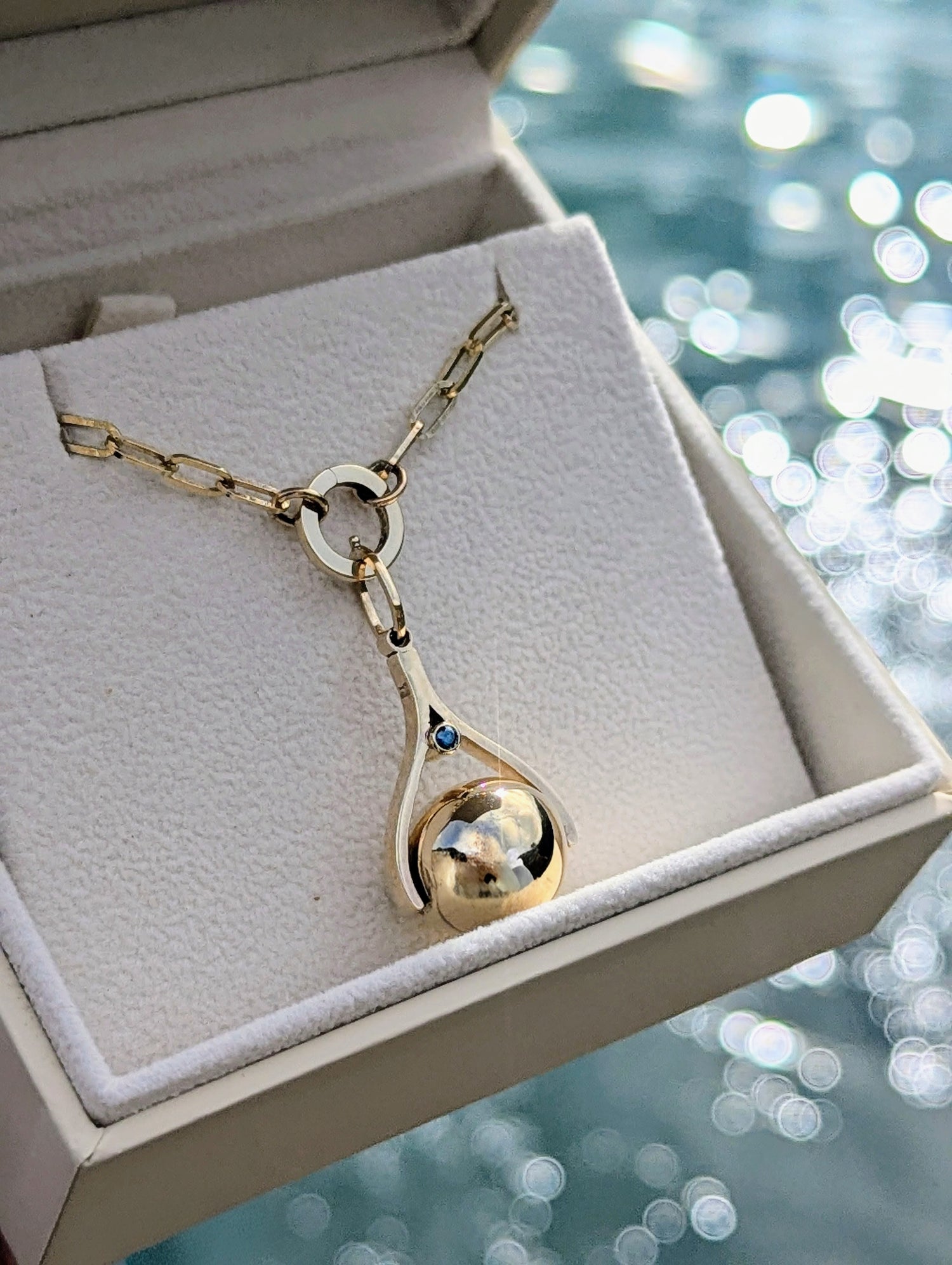 Gold necklace with a spinning pendant in an open jewellery box by a shimmering ocean water.