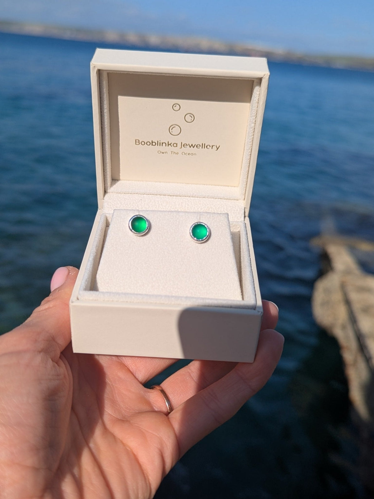 Pair of bright green sea glass stud earrings in sterling silver, held up in a jewellery box against ocean background.