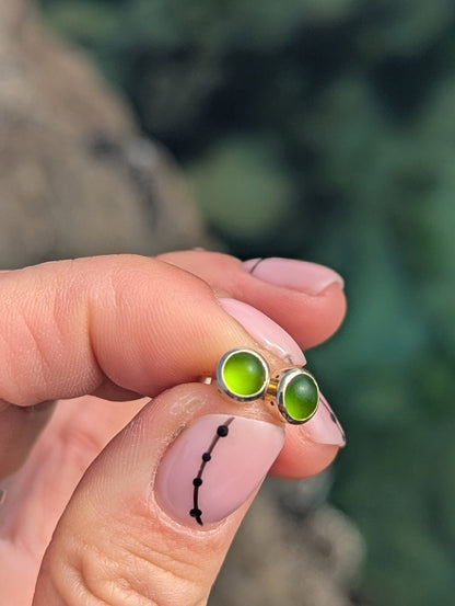 Close-up of a green sea glass stud earring held between the fingers, showing the gold setting and bright green colour.