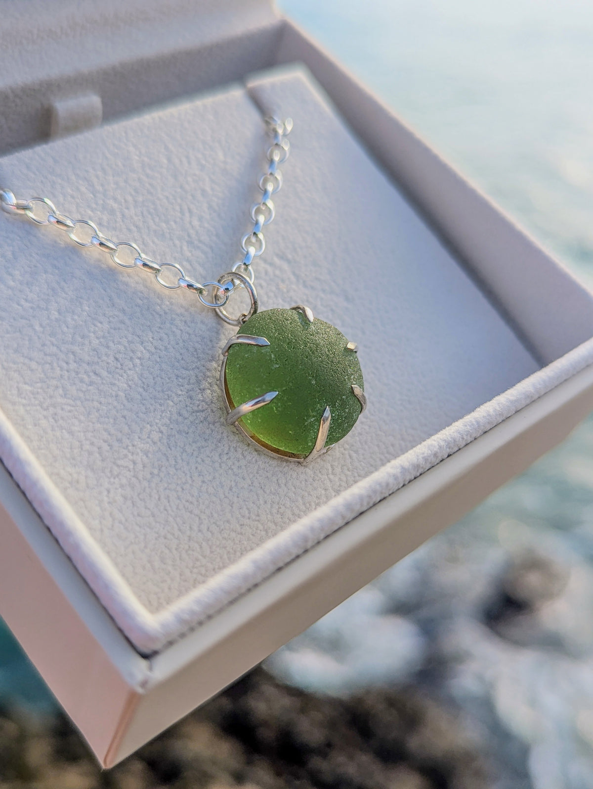A green sea glass pendant necklace in a silver setting on a silver chain, displayed inside an open white jewellery box with a blurred ocean background.