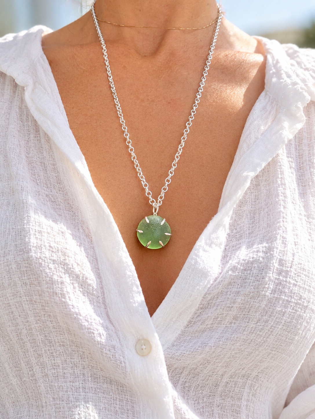 A close-up of a person wearing a silver chain necklace with a round green sea glass pendant, styled with a white textured blouse.