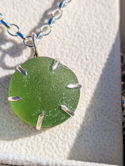 A close-up of a sea glass pendant necklace with a round green sea glass set in silver, hanging from a silver link chain on a textured white surface.
