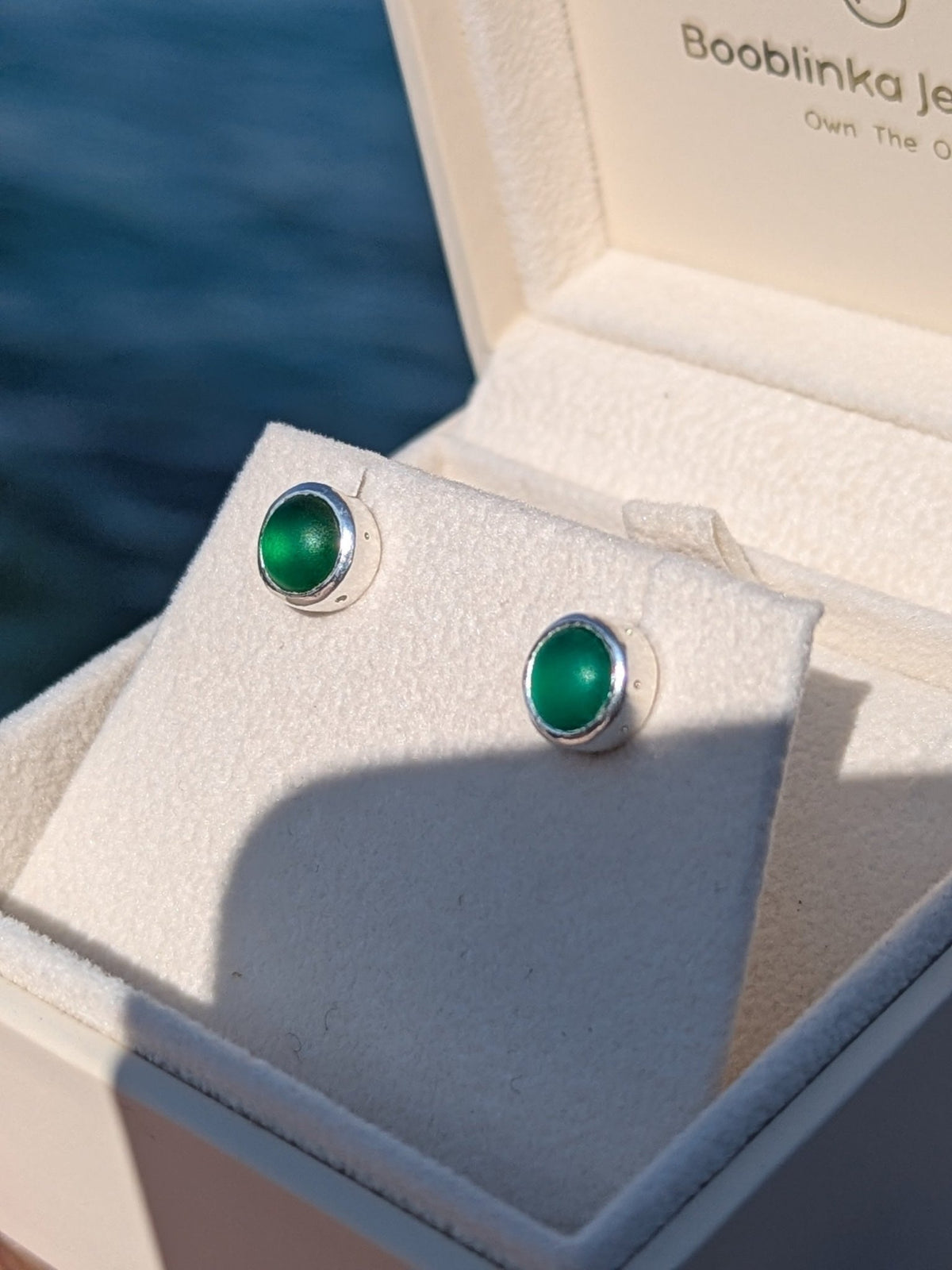 Close-up of two round green sea glass studs in silver settings, displayed inside a jewellery box against ocean background.