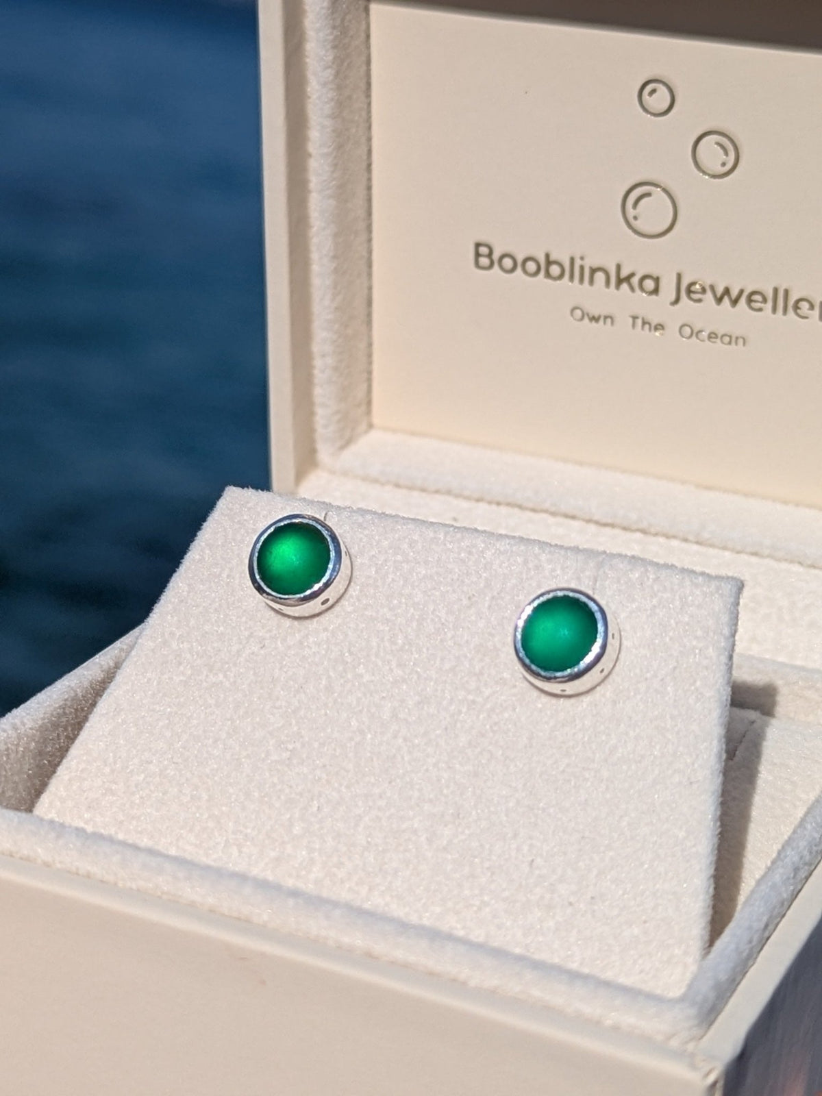 Green sea glass stud earrings set in sterling silver, displayed in a white Booblinka Jewellery box with ocean background.