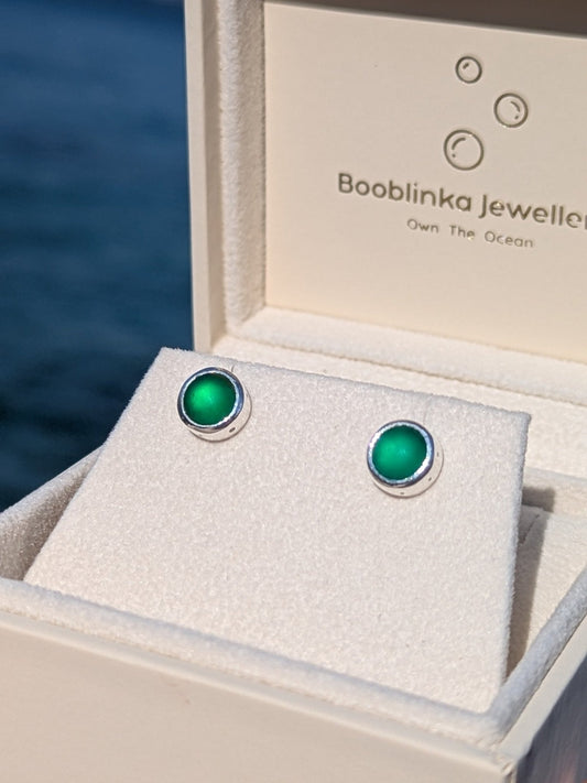 Green sea glass stud earrings set in sterling silver, displayed in a white Booblinka Jewellery box with ocean background.