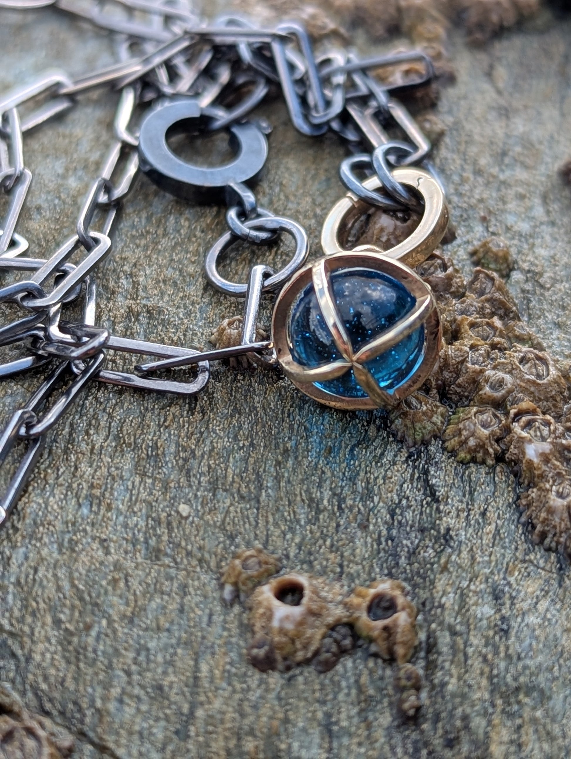 Handmade Swiss Blue Topaz sphere necklace with a solid 9ct gold cage, 9ct gold hinged charm holder and black rhodium sterling silver chain displayed on weathered coastal rocks in Cornwall.